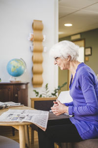 Side view of senor woman smiling while reading newspaper at table in nursing home