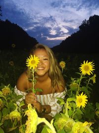 Portrait of young woman holding sunflower on field against sky at dusk