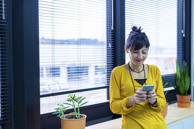 Happy businesswoman using smart phone against window in office