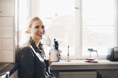 Happy woman with coffee cup against window in kitchen at home