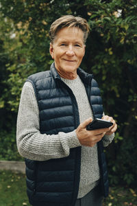 Portrait of smiling senior man holding smart phone while standing at back yard