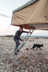 Side view full body of male hiker climbing up on ladder to camping tent on roof of car during hiking journey with dog in vast valley