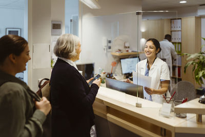 Patients talking to female receptionist through transparent shield in clinic