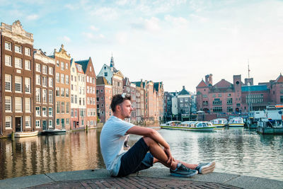 Side view of man sitting by canal against buildings in city