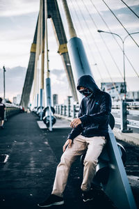 Full length of man sitting by bridge against sky