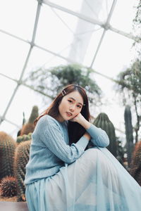 Portrait of young woman sitting against plants in greenhouse