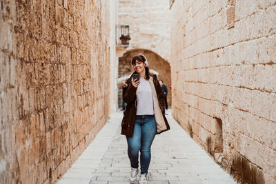 Full length of woman wearing headphones walking in alley amidst building