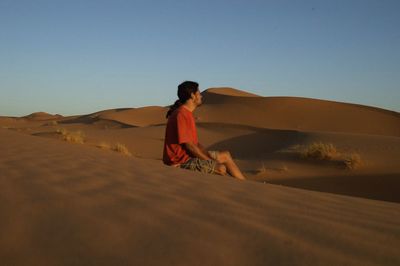 Man sitting on sand dune against clear sky at desert