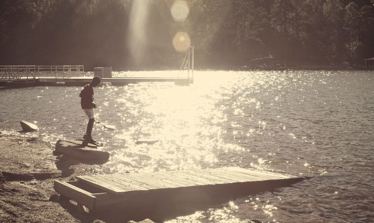 REAR VIEW OF MAN STANDING BY LAKE AGAINST TREES