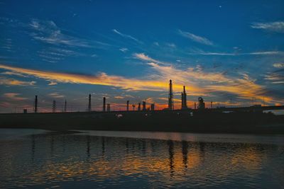 Silhouette factory by river against sky during sunset