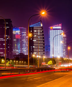 Light trails on road by illuminated buildings against sky at night