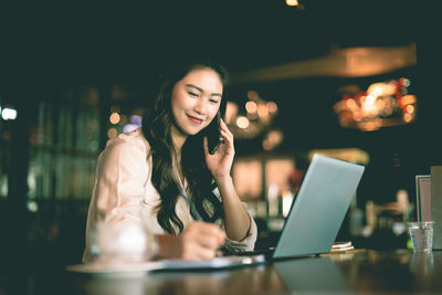 Smiling woman talking on phone while using laptop at cafe