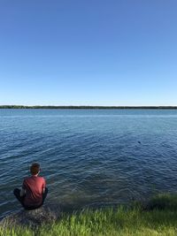 Man sitting on lake against clear blue sky