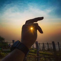 Optical illusion of hand holding sun against sky during sunset