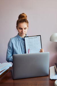Cute girl holding clipboard standing by laptop at home