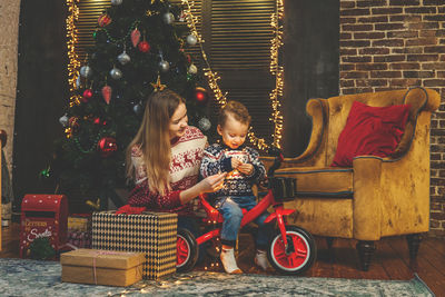 Mother and son with gift sitting on chair against christmas tree