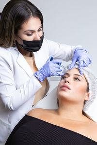 Graceful young female client with long blond hair sitting on medical chair while cosmetologist in latex gloves making rejuvenating facial injections procedure in beauty center