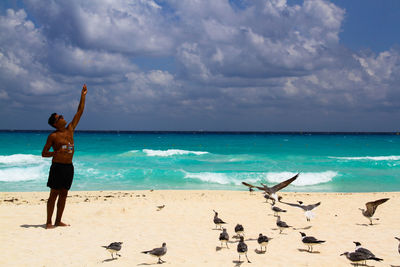 Shirtless mature man standing at beach against cloudy sky