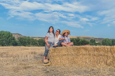 Mother and daughter with hay bale on landscape against blue sky