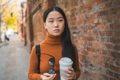 Beautiful woman holding sunglasses and disposable cup while standing against brick wall