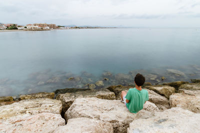 Rear view of boy looking away while sitting on rock by sea