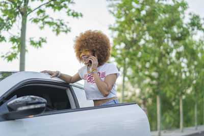 Woman with afro hair talking on the phone next to her white car