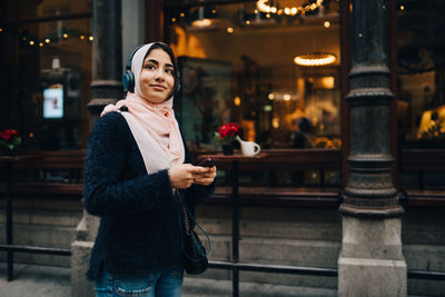 Teenage girl listening to headphones holding smart phone while looking away in city