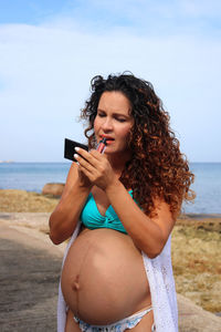 Pregnant woman putting lipstick on her lips at seaside 