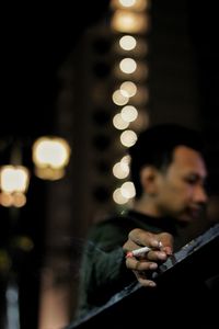 Side view of young man holding cigarette against defocused lights at night