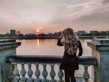 Rear view of woman standing by railing against cityscape during sunset