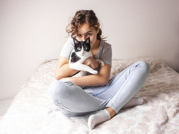 Portrait of girl with cat sitting on bed against wall at home