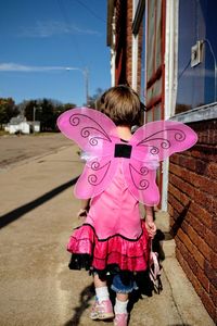 Rear view of girl wearing pink wings while walking on footpath by building in city