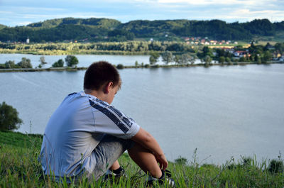 Rear view of boy sitting on lake against sky