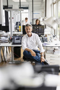 Mature businessman sitting on chair with colleague standing in background at open plan office