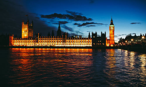 View of illuminated tower bridge over river by buildings in city at night