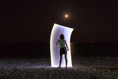 Rear view of man standing against illuminated sea at night