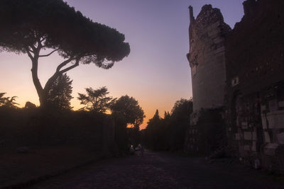 Road amidst silhouette trees and buildings against sky at sunset