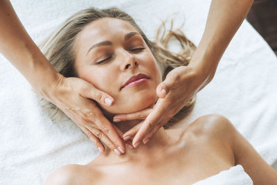 Attractive blonde young woman on couch with white linen enjoys hand facial massage in the spa salon