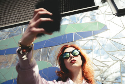 Low angle view of redhead young woman taking selfie with mobile phone against mosaic wall