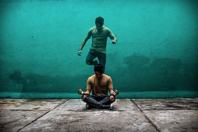 Man jumping by friend sitting on footpath against wall