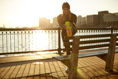 A backlit woman stretches pre-run on a park bench.