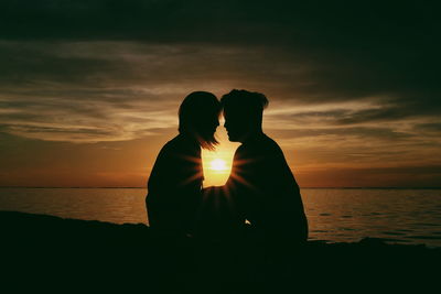 Silhouette couple sitting on beach against sky during sunset