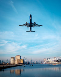 Low angle view of airplane flying over river against blue sky in city