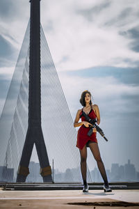 Portrait of young woman standing against sky. woman in red aiming a gun