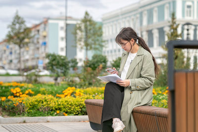 A girl sits on a bench on the street with documents in her hands and using her phone