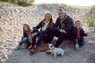 Family with a son and daughter and a dog sit on the sand in autumn in leather jackets