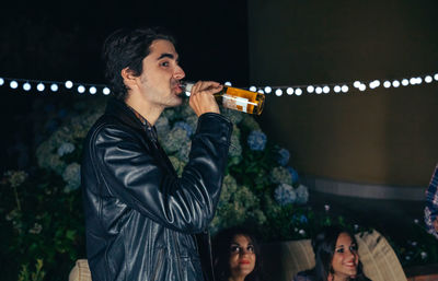Side view of young man drinking beer while standing at patio during night