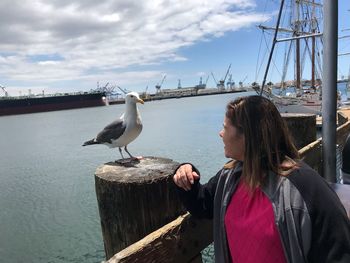 Woman looking at seagulls perching on wooden post against sky