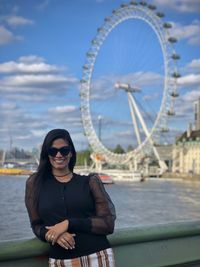 Portrait of woman wearing sunglasses against ferris wheel by river in city