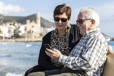 Man using mobile phone by woman sitting against sea and sky